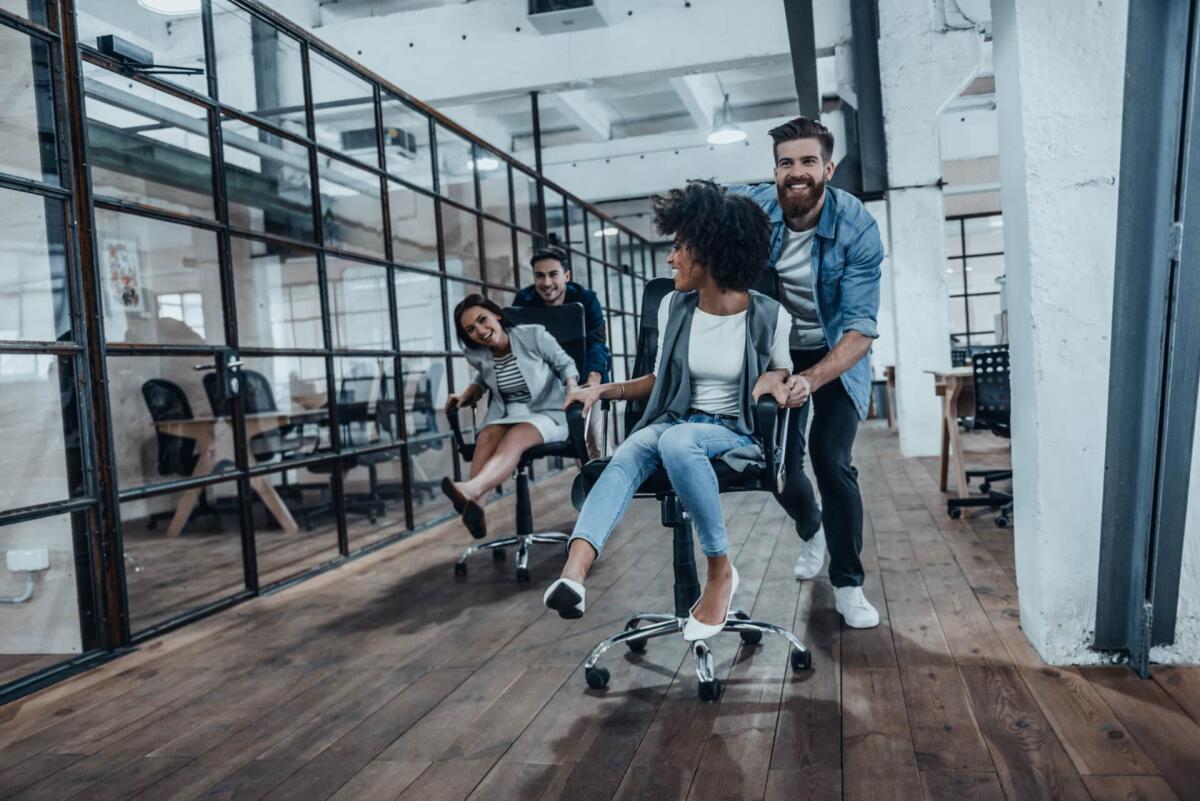 Sedie da ufficio ergonomiche: modelli e prezzi - Unstoppable team! Four young cheerful business people in smart casual wear having fun while racing on office chairs and smiling