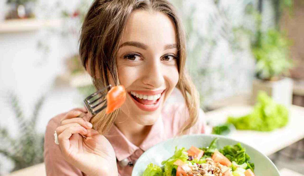 Cibo e felicità: quali sono gli alimenti che aiutano a combattere? - Immagine di donna che sorride e mangia (Canva Foto) - www.statodonna.it